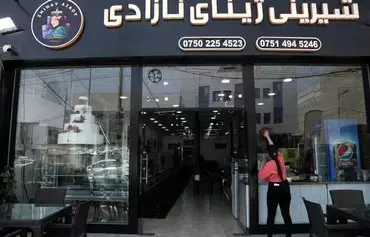 On July 15, 2023, an Iraqi Kurdish woman wipes the window of a confectionary store and café in Erbil, Iraq. The store is named after Mahsa Amini, a young Iranian Kurdish woman whose September 2022 death in custody of the Iranian regime's morality police sparked months-long nationwide anti-regime protests in Iran. [Safin HAMID/AFP]