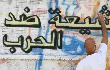 مردی با استفاده از اسپری روی دیواری در لبنان یک طرح گرافیتی را رسم می‌کند که روی آن نوشته شده است: «شیعیان مخالف جنگ هستند.» [صفحهٔ فیسبوک شیعیان علیه جنگ]