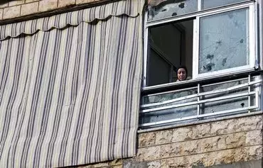 A woman in a building near the site of the January 2 drone strike that killed Hamas deputy leader Saleh al-Aruri in the southern suburb of Beirut, looks out of a window that was damaged in the blast, on January 3. [Anwar Amro/AFP]